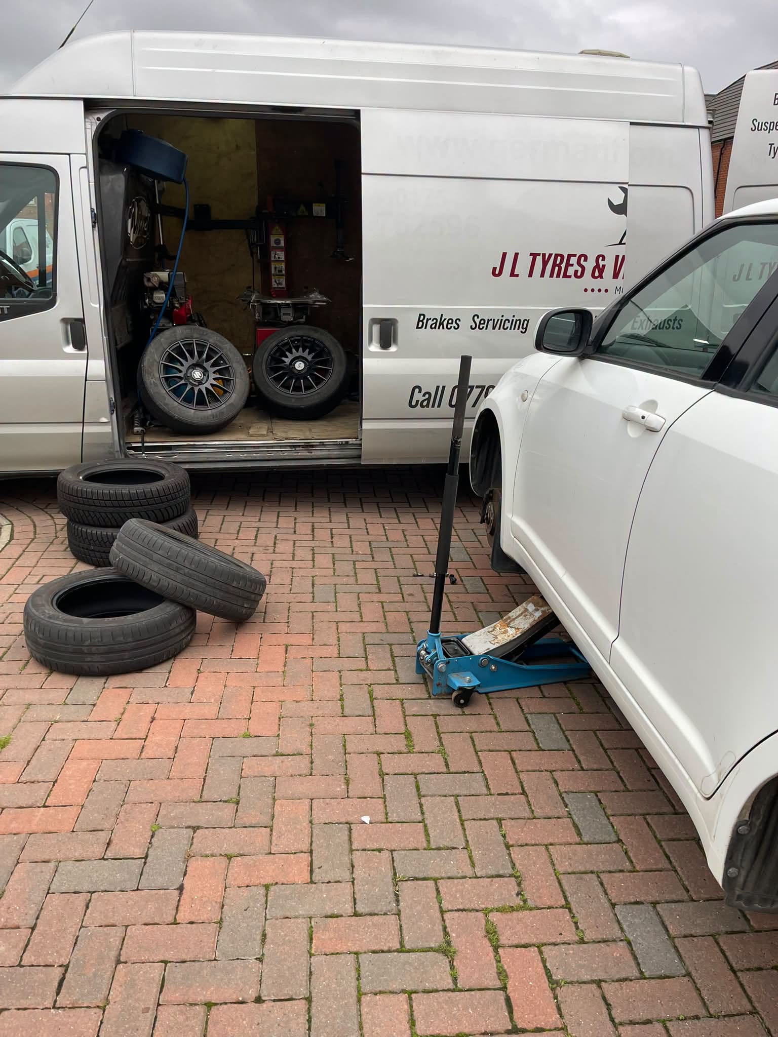 The J L Tyres van with wheels inside and a white car on a jack on block paving